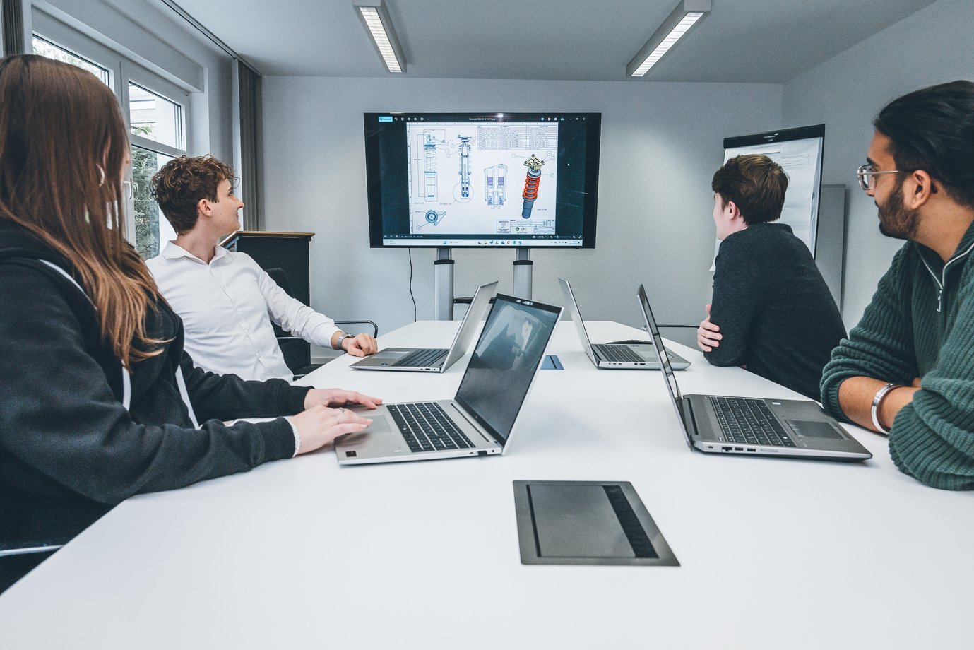 Personen sitzen mit Laptops in einem Besprechungsraum und schauen auf einen großen Bildschirm mit technischen Darstellungen.