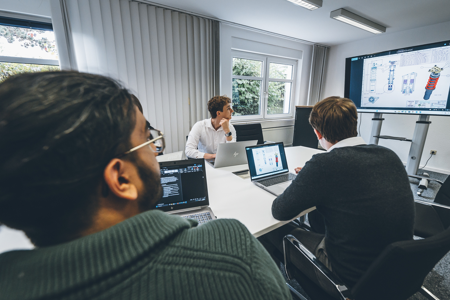 Personen sitzen mit Laptops in einem Besprechungsraum und schauen auf einen großen Bildschirm mit technischen Darstellungen.